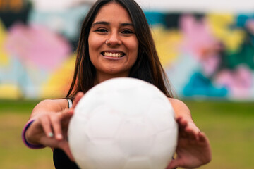 Naklejka premium View of a beautiful brunette female soccer player posing, holding and showing a soccer ball. On a green grass outdoors, with a colorful background. Sportswear. Sports and wellness advertising.
