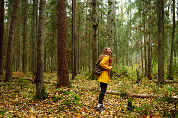 A woman in a yellow rain jacket explores a lush forest during a cool autumn day, surrounded by tall...