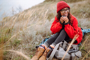 Young woman enjoying a warm drink while sitting on a blanket in a grassy field during a cool autumn day.  Feeling harmony, relax, personal fulfillment. Camping.