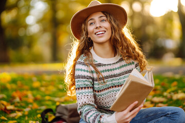A young woman enjoys reading by a serene lake surrounded by autumn leaves in a park during sunset....