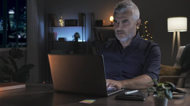 mature man sits at the home office desk working late using laptop computer  at night,middle aged male uses notebook in the dark room