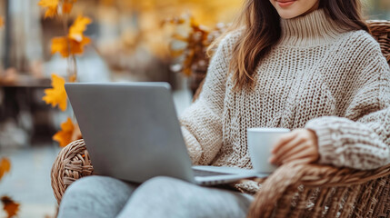 Beautiful woman working on a laptop computer while sitting in a wicker chair at a outdoors autumn cafe with cozy fall street atmosphere , a coffee cup near her hand