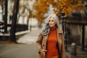 beautiful, elegant senior woman in a orange sweater and brown coat, with long grey hair, walking on the autumn street and looking at the camera. Concept for business success, fashion and aging