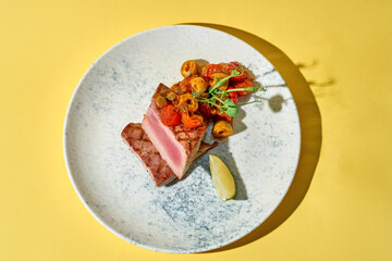 seared tuna steak is presented on against a yellow background. The tuna is sliced to reveal a beautiful pink center, and is topped with a medley of roasted cherry tomatoes and capers