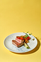 seared tuna steak is presented on against a yellow background. The tuna is sliced to reveal a beautiful pink center, and is topped with a medley of roasted cherry tomatoes and capers