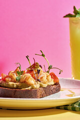 A gourmet open-faced sandwich on a white plate is adorned with grilled shrimp, a vibrant tomato salsa, and a creamy hummus spread.