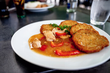 Chicken piece in red pepper sauce, small potato pancake on plate, glass of water on table.