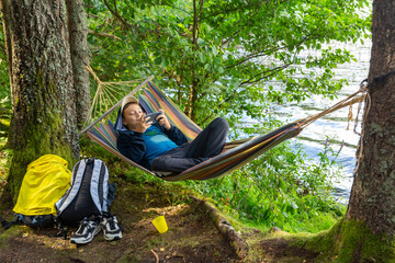 A teenage boy is relaxing in a hammock on the shore of a forest lake and playing with a smartphone.