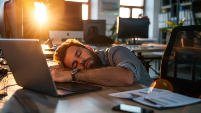 A young professional leans on his desk, fast asleep, surrounded by office equipment and paperwork as the warm light of sunset filters through the windows