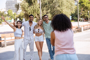 Four friends pose for a photo in a sunny park, smiling and waving while their friend captures the moment, with trees and benches in the background. 