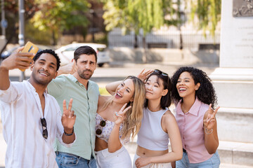 A happy group of five diverse friends capturing a selfie outdoors, smiling and posing with peace signs on a sunny day, enjoying a fun moment together. 