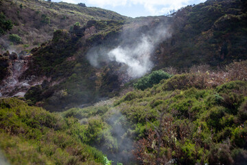 Sulfurous steam rises from the rocky landscape of Furnas de Enxofre, Terceira Island, Azores, Portugal. A unique geothermal site surrounded by lush green vegetation and rugged terrain.