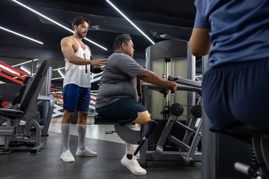 Asian man with prosthetic leg from amputation from diabetes doing weight training workout exercise machine at gym with professional trainer for handicap and disability people and recovery