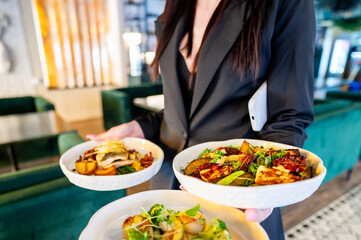 person holding three plates of food, showcasing a variety of dishes in a restaurant setting. Ideal for themes related to dining, hospitality, and culinary experiences. Bright and inviting atmosphere.