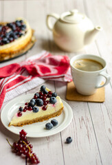 Cheesecake with berries on white plate and coffee in white mug on wood table with red towel