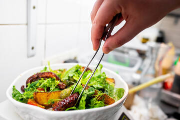 A close-up of a fresh, vibrant salad with mixed greens, ripe tomatoes, and various vegetables being tossed with tongs in a white bowl, suggesting a healthy lifestyle.