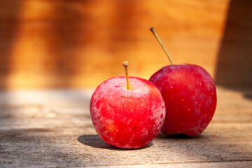 Two small, vibrant red crabapples, each with a stem, sitting on a textured wooden surface with warm sunlight and shadows. A simple and natural still life