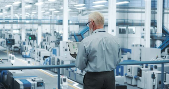 Middle Aged Engineer Monitoring and Analyzing Conditions at an Automated Modern Electronics Factory with Mobility Robots Working with the Help of Artificial Intelligence Software. Back View Footage