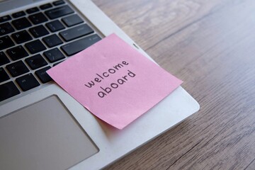 A close-up of a laptop keyboard with a pink sticky note attached with text “Welcome aboard”,...