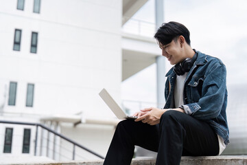 Smiling Student Using Laptop Outdoors on Campus, Wearing Headphones and Casual Clothing, Engaged in Online Learning or Studying