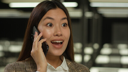 Amazed wonder surprised Asian girl student indoors listening good news mobile phone call excited happy Korean business woman Japanese businesswoman Chinese female in office talk smartphone achieve