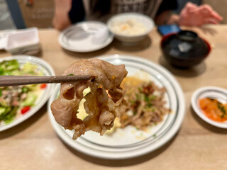 chopsticks holding a slice of tender, cooked meat with a blurred background of a Japanese meal. The focus is on the texture and juiciness of the meat, showcasing the delicious 