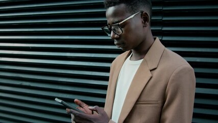 Tilt up shot of Black businessman dressed formally replying to messages using smartphone while waiting for colleague to pick him up near office building