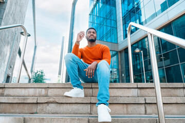 A Man in an Orange Sweater Relaxes on Steps Outside a Modern City Building Under a Bright Sky