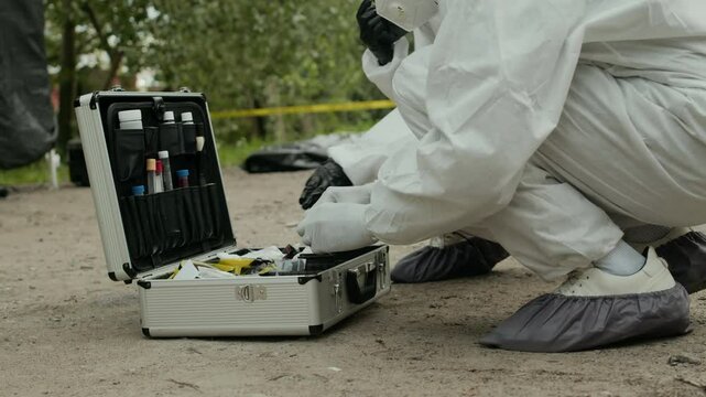 Team of crime scene investigators gathering forensic evidence on ground near dead body lying under plastic bag