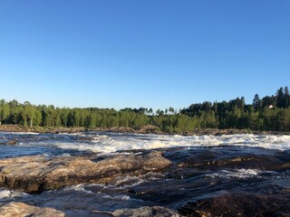 A serene landscape of a rushing river surrounded by lush forest and a clear blue sky, symbolizing the power and tranquility of nature.