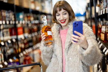 Woman chooses strong alcohol while standing with bottle and phone in supermarket. Buying cognac...