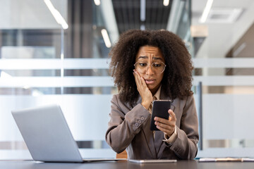 Shocked and scared young African American woman is in the office, sitting at the desk and looking...