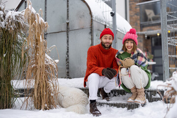 Young couple in sweaters hats and gloves sit and warm together on a porch of their house, spending...