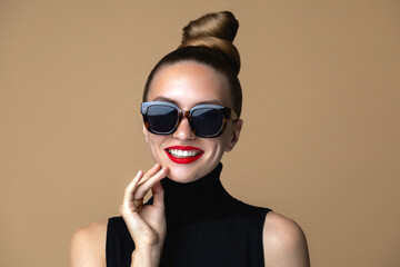 A stylish woman with a chic top bun hairstyle wearing black sunglasses, red lipstick and a black turtleneck smiles confidently against a beige background. Eye wear fashion