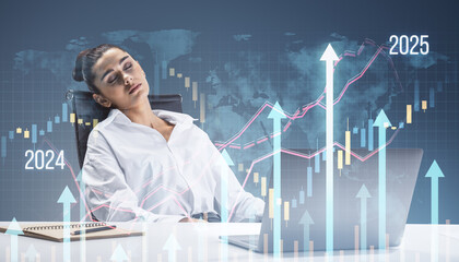 Attractive sleepy young woman sitting at desk with laptop, supplies and growing business chart grid hologram and map on blurry background. Financial growth and fintech concept. Double exposure.