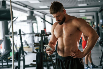 With smartphone, taking a break. Strong muscular man is working out in the gym