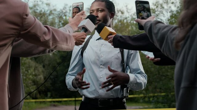Medium shot of Black male police detective in workwear giving interview to press on -location during crime scene investigation