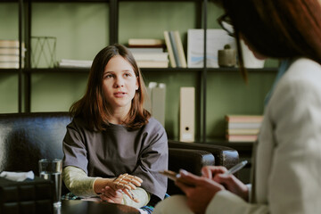 Young girl sitting opposite healthcare professional in office, discussing health concerns and holding hands together with a thoughtful expression