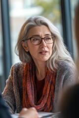 A woman with glasses and a scarf is sitting at a table. She is looking at something and she is deep in thought
