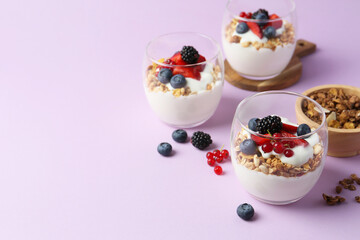 Tasty yogurt with fresh berries and granola in glasses on violet background, space for text