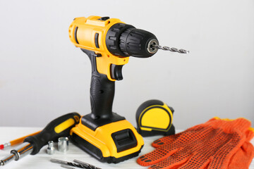 Cordless electric drill, gloves, bits, screwdriver and measuring tape on white wooden table