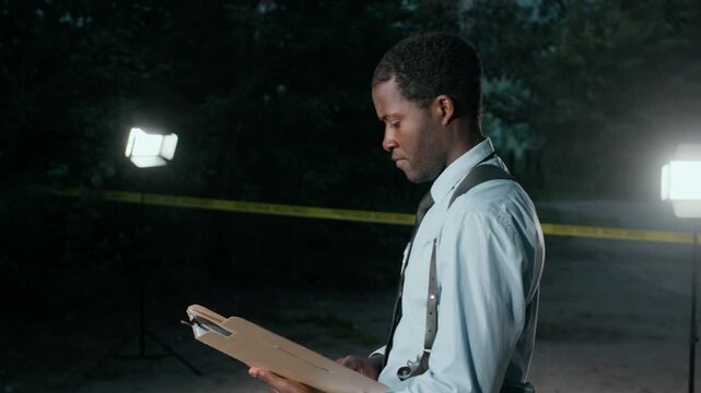 Medium shot of African American male police detective investigating criminal case at night, going out of tent set up right at crime scene and looking at documents in folder