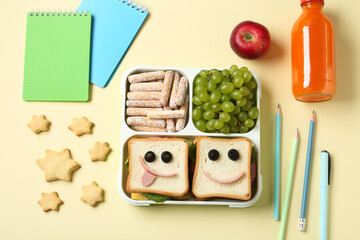 Sandwiches with smiley faces in lunch box, bottle of juice and stationery on yellow table, flat lay