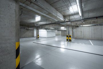 Underground parking garage with concrete pillars, empty parking spaces, yellow and black safety markings, illuminated with modern infrastructure and installations, located in an urban setting.