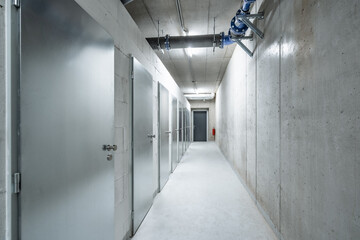 A long, narrow, concrete corridor with metallic doors, industrial lighting, and no visible people, leading towards a closed exit door at the far end.