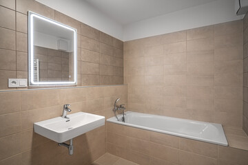 Modern bathroom with a bathtub, illuminated mirror, and a sink, featuring neutral beige tiles, sleek fixtures, and minimalist design.