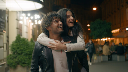 Fototapeta premium Indian Arabian couple love guy holding girl on back piggyback woman man boyfriend girlfriend walking night evening city looking at beautiful urban architecture pointing excited tourist travel dating