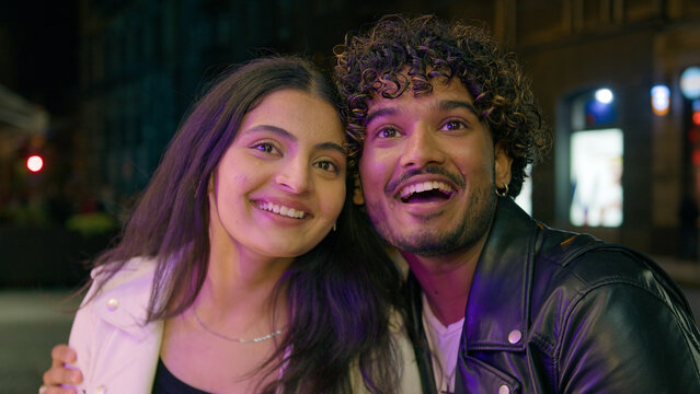 Arabian Indian happy couple in love in evening city dating urban street night looking pointing at showcase talking laughing smiling discount choosing presents look at digital screen reflection cinema