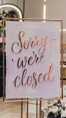 Stylish "Sorry We're Closed" sign in shiny rose gold script on a pastel lavender background, framed in an elegant boutique setting. Refined floral accents and soft lighting enhance the luxurious