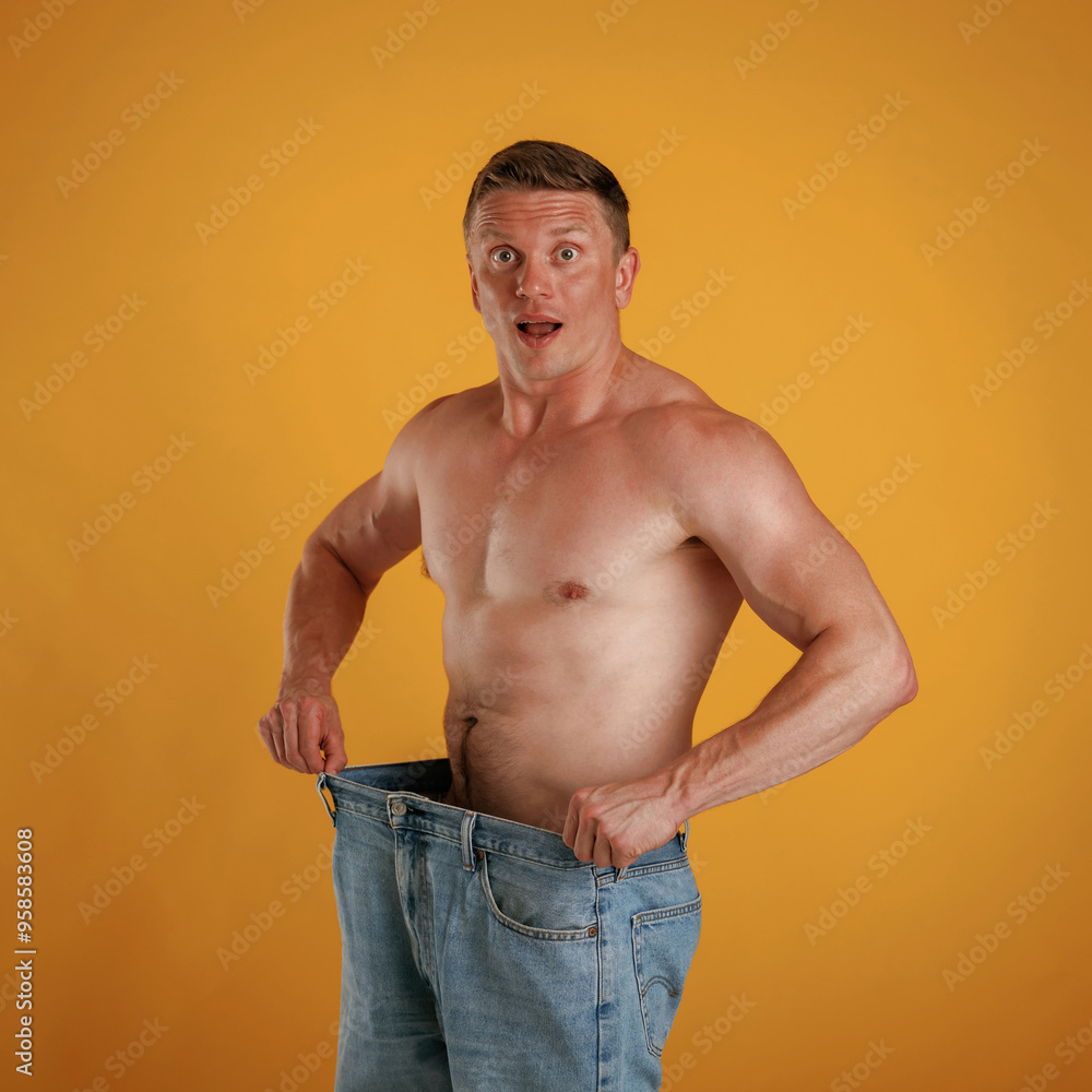 Wall mural Weight loss, shocked by results, big and huge pants or jeans. Man is against background in the studio